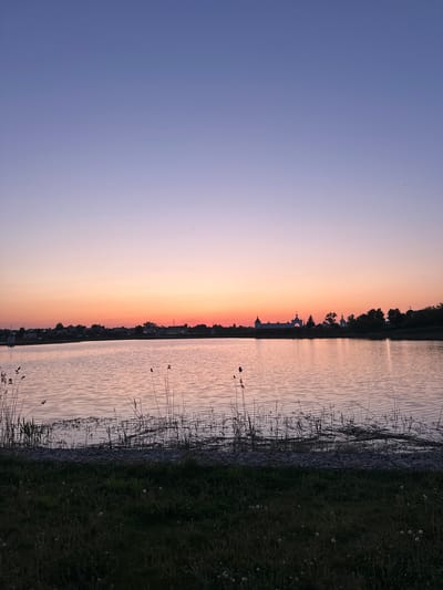 The image depicts a tranquil landscape at sunset in what appears to be Diveyevo, Russia. The sky transitions from deep blue on the left to hues of orange and pink near the horizon. A body of water, possibly a lake or a wide river, reflects the sunset colors. Along the shoreline, vegetation, including grasses and reeds, is visible. In the distance, the silhouetted outline of a building with domes suggests a church or monastery, a typical architectural feature of Russian Orthodox religious sites, further suggesting the location is likely Diveyevo, which is known for its prominent monastery. The lighting indicates a calm, late-day atmosphere.