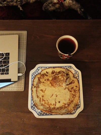 The image depicts a top-down view of a wooden table. On the table, there is a laptop computer with a white cable plugged into it, a cup of dark liquid, and a plate of crepes. The plate has a blue and white checkered pattern on its rim. The laptop is positioned on a gray cutting mat with grid lines. In the background, there is a dark, textured surface, possibly a rug or blanket, with hints of red and white. The lighting is dim, suggesting an indoor setting. There are no people visible in the image. The scene is set on a wooden table, likely in a home or cafe. Given the provided location of Шарпілаўскі сельскі Савет, Belarus, this scene is likely taking place within a residential or communal space in that area.