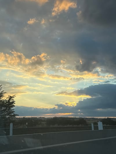 The image depicts a dramatic sky with a sunset casting golden light through dark clouds. The scene appears to be taken from a vehicle traveling along a highway, as indicated by the concrete barrier in the foreground and the landscape stretching towards the horizon. The landscape features a line of trees and vegetation, with some distant structures visible. There are no people or specific identifiable landmarks of Bad Dürrheim in the image. The overall impression is a moment captured during a scenic drive with an impressive cloud formation.