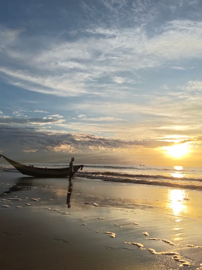 The image depicts a beach scene at sunrise in Uleekareung, Indonesia. A single person, silhouetted and wearing a light-colored garment, stands beside a long, narrow boat on the wet sand. The boat is positioned parallel to the shoreline, facing towards the water. Gentle waves are washing ashore, creating a reflection of the sky and the sun on the wet sand. The sky is filled with scattered clouds, with the sun just breaking through on the right side of the frame, casting a warm, golden glow across the water and the beach. The overall atmosphere is serene, capturing the quiet moments of early morning at the coast.