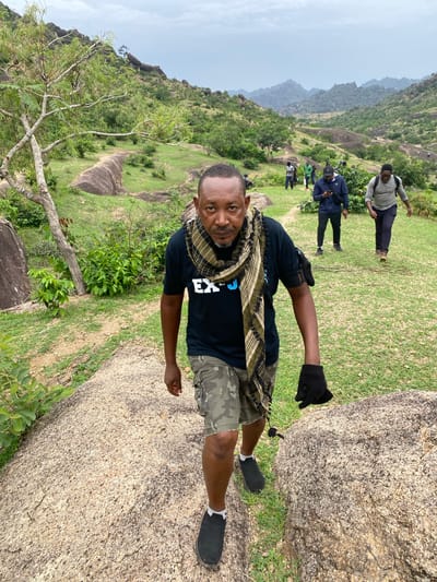 The image shows a man in a black t-shirt, camouflage shorts, and black shoes standing on a large rock formation. He has a patterned scarf around his neck and a black glove on one hand. In the background, there is a landscape with other people, grass, trees, and more rock formations. The location appears to be a hilly or mountainous area. The image shows the location of Ganawuri, Nigeria.