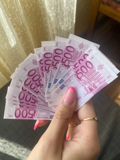 The scene, located in Kipilovo, Bulgaria, presents a close-up view of a human hand holding a fan of multiple 500 Euro banknotes. The hand, positioned in the lower right and center, exhibits manicured nails with a light pink base and vibrant pink tips in a French manicure style. A thin, gold-toned ring is worn on the ring finger. At least eleven 500 Euro banknotes, predominantly purple in hue, are fanned radially, clearly displaying the "500" denomination, "EURO" text, and characteristic architectural designs. The background on the left consists of a light-colored, vertically textured fabric, likely a curtain, illuminated by natural light. The background on the right is darker and out of focus, suggesting an indoor environment with indistinct furniture and textiles.