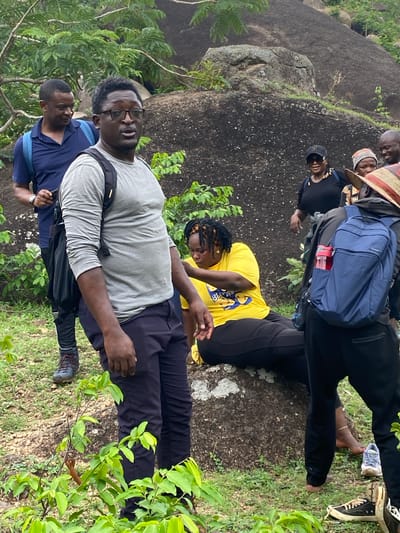 A group of people stand near a large rock formation, likely at the Ri-Yom Rock Formation in Ganawuri, Nigeria. Some of the people are wearing backpacks and hiking shoes. There are bushes and trees nearby. One person is wearing a hat.