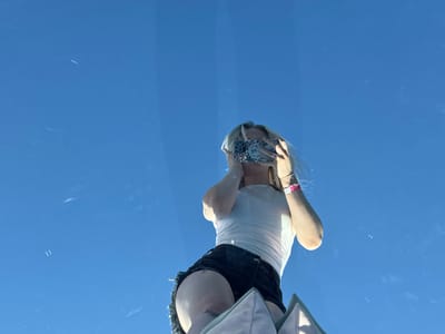 A low-angle shot captures a person with blond hair wearing a white tank top and black shorts. They are holding a decorated phone up to their face as if taking a picture, obscuring their facial features. The backdrop is a bright blue sky, suggesting an outdoor setting. The person's legs and part of a gray object are visible in the lower portion of the frame. Streaks and marks on the surface create a sense of depth.