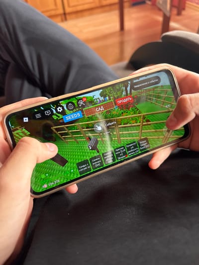 The image shows a person's hands holding a smartphone, playing a game. The game appears to be a farming or gardening simulation, with on-screen buttons and graphics depicting crops, tools, and gameplay elements. The phone is held in landscape orientation. The background is out of focus, but appears to be an indoor setting with wooden furniture and flooring, possibly a living room or dining room. There is no visible indication of the city of Ветлужский, Russia.