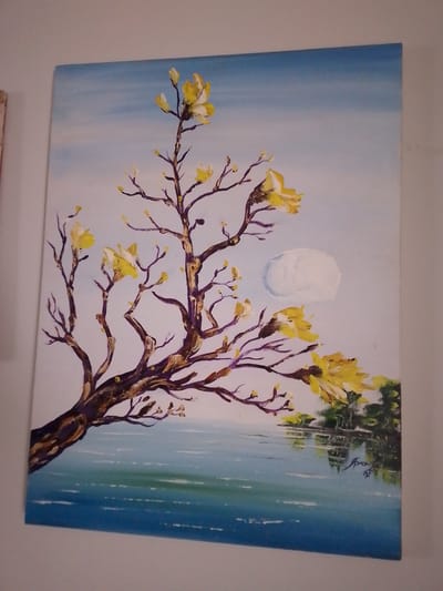 A photograph captures an oil or acrylic painting displayed on a light-colored wall. The painting depicts a serene landscape, possibly located in or inspired by the environment of Campo Grande, Brazil. A large, gnarled tree with textured dark brown branches and vibrant yellow blossoms or buds extends from the bottom-left foreground, diagonally across the center, towards the upper-right. The sky transitions from light blue at the top to a paler hue towards the horizon. A distinct, impasto-rendered white circular object, resembling a moon or stylized cloud, is positioned in the upper-right sky. The lower half of the painting illustrates a calm body of water, rendered in blue and green tones, reflecting a distant shoreline with small clusters of green foliage on the right. A dark, calligraphic artist's signature is visible in the lower-right area, within the water's reflection. No people are present, and no direct actions are depicted.