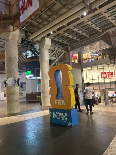 The image shows an indoor scene, likely a shopping mall, with a large, abstract sculpture in the foreground. The sculpture is yellow and blue and has the word "FIFA" visible on it. Next to the sculpture is a blue box with text that includes "KALLA" and "A PART OF KALLA". Two people are walking in the background, wearing casual clothing. One person is wearing a white t-shirt and dark pants, and the other is wearing a checkered shirt and dark pants. Behind them, there are storefronts, including one with the "UNIQLO" logo. The lighting suggests it is daytime, and the setting appears to be a modern, well-lit interior space. The overall atmosphere seems relaxed and ordinary, with no specific event indicated. The architectural elements of the building, such as exposed beams and pipes, are also visible.