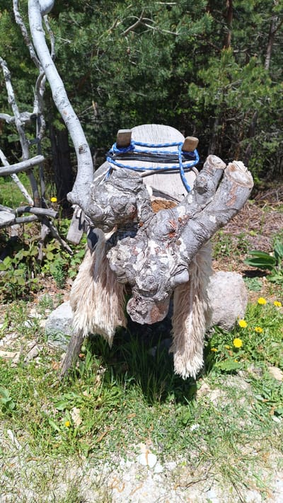 The image shows a wooden sculpture of an animal, possibly a yak, in an outdoor setting. The sculpture is made from tree branches and logs, with a rough-hewn appearance. It has a saddle-like structure on its back, secured with blue rope. The sculpture is covered with a layer of shaggy material that looks like sheep’s wool. The surrounding environment consists of grass, dandelions, and trees.

There is no additional data to pinpoint the sculpture's exact location in Sini Vrah, Bulgaria.