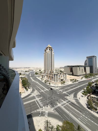 The image, viewed upright, presents a high-angle panoramic view of a modern urban landscape in Lusail, Qatar, under a clear and bright blue sky.
In the immediate foreground, the curving white edge of a building or balcony structure occupies the lower left corner. The midground is dominated by a prominent, slender high-rise building characterized by an ornate, possibly golden, pointed roof or spire. To its right, a complex network of multi-lane roads, highways, and intersections unfolds. The roads are meticulously planned with crisp white lane markings, directional arrows, and pedestrian crosswalks. Traffic lights and streetlights line the roadways, but the roads themselves appear largely empty, with only one white car visible, suggesting minimal activity or traffic.
Further into the scene, several other modern buildings with distinct architectural designs are visible, including one with an intricate, geometric patterned façade. Palm trees dot the landscape along the roads and between buildings. The urban environment extends towards a hazy horizon in the distance, showcasing extensive development. The clear sky and bright light indicate it is daytime. There is no visible text in the image.