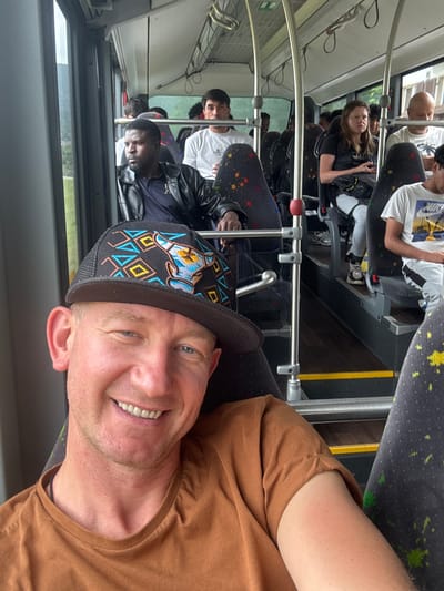 A man with a brown t-shirt and a patterned baseball cap smiles at the camera while riding on a bus. He is in the foreground, with other passengers visible in the background. The bus appears to be traveling through a wooded area, likely in the vicinity of Reconvilier, Switzerland, given the typical scenery in the region. The interior of the bus shows grey seats with colorful speckles, metal poles for support, and large windows offering views of the outside. The man's cap has a detailed design with blue, yellow, and black geometric patterns and a stylized animal head. The lighting suggests it is daytime.