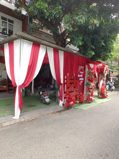 The image depicts a decorated building entrance in Phú Xuân Ward, Vietnam, viewed from the street level. A corrugated metal awning extends from a white multi-story building, beneath which elaborate red and white fabric drapes create a festive enclosure over the sidewalk. The drapes are tied back to form an arched entrance on the left, revealing interior seating. The sidewalk area directly beneath the awning is covered with green artificial turf.

To the right of the fabric entrance, a large red "Double Happiness" (囍) symbol stands prominently, surrounded at its base by clusters of red artificial flowers. Adjacent to this, a small pink oval sign with Vietnamese text is affixed. Further to the right, two decorated vertical pillars, adorned with red, orange, and green artificial flowers and foliage, frame another entrance or display area.

A grey scooter with a visible license plate ("75-F1 376.26") is parked on the green turf, facing slightly left. A large tree with dense green foliage grows immediately above the awning, its trunk partially visible. The foreground consists of a grey asphalt road with a concrete curb, on which scattered leaves and debris are visible. In the distance on the right, another scooter is partially visible on the street. No individuals are present in the immediate scene. The lighting suggests daytime conditions.
