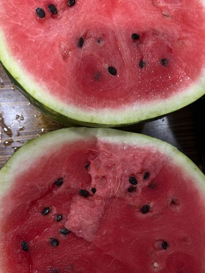 The image shows a close-up, overhead view of two halves of a sliced watermelon. The watermelons are a vibrant pink-red flesh with visible black seeds and a light green rind. The surface the watermelons are resting on appears to be a wooden table with some water droplets scattered on it, suggesting a recent cut or moisture. The lighting is bright, highlighting the glistening texture of the watermelon flesh. There are no people or explicit text visible in the image. The scene is focused entirely on the fresh, juicy appearance of the watermelon halves. The context provided places the image in Ильчимбетово, Russia, but this location is not visually discernible from the image itself.