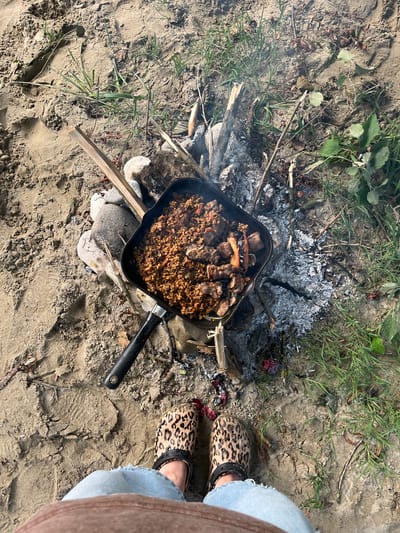 A top-down, medium shot captures a meal being prepared outdoors on a sandy patch of ground amidst dry grass and scattered leaves. The focal point is a black frying pan filled with a grain and meat dish, placed over a small, smoldering campfire. Embers glow beneath the pan, and wisps of smoke rise from the food.

To the right of the pan, a person's legs are visible, clad in light blue jeans with a ripped knee, and wearing leopard-print clogs. A beige fabric edge, likely from a garment, runs down the side of the frame.

The ground is composed of sand and dirt, with patches of green grass and dried leaves scattered throughout. Small stones and pieces of wood are also present, some forming part of the makeshift campfire. A single, split wooden stick juts out from the sand in the lower right quadrant.

The scene suggests a casual outdoor meal, possibly a picnic or camping trip. The lighting indicates it is daytime, with no strong shadows suggesting it is not midday. The overall mood is relaxed and rustic. No visible text is present in the image. The context provided indicates the location as Nalchik, Russia.