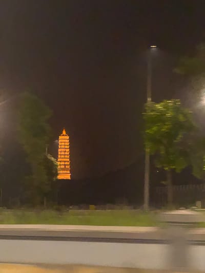 A night time photograph captures a pagoda, illuminated by orange lights, in the distance. The pagoda has a stepped, multi-tiered structure, with a pointed roof. Surrounding the pagoda are dark trees and foliage. In the foreground, a street light with an illuminated head stands to the right of the pagoda, also casting a warm glow. A road with a grey barrier runs horizontally across the bottom of the image, with a strip of green grass and more foliage visible behind it. The sky is dark, suggesting it is late evening or night. The overall scene is one of an urban landscape at night, with a prominent architectural landmark. This pagoda is likely the Trấn Quốc Pagoda, a historical and cultural site located in the Tây Hồ District of Hanoi, Vietnam, known for its beauty and spiritual significance.
