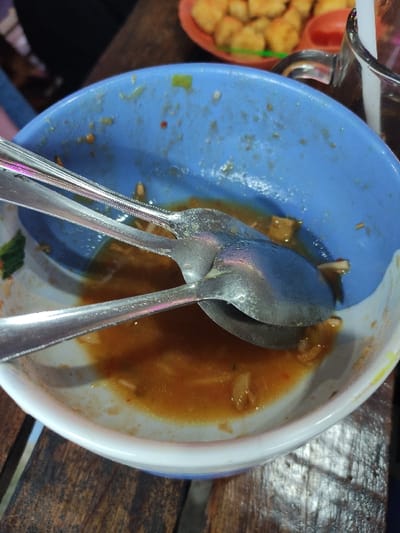 The image captures a close-up, overhead view of a blue bowl with a white rim, mostly empty but containing remnants of a reddish-orange sauce and a few small food particles. Two silver-colored metal spoons rest inside the bowl, partially covered by the remaining liquid.
To the upper left, a portion of a clear glass or cup is visible. Further left, part of a red or orange basket or container can be seen, filled with numerous small, golden-brown, cuboid-shaped fried items, likely fried tofu or tempeh. All items are situated on a dark wooden table surface, which shows visible grain.
The scene depicts the aftermath of a meal or snack, indicated by the nearly empty bowl with leftovers. No people are visible, nor is any text. The setting is a casual environment, consistent with a mealtime in Kecamatan Pandaan, Indonesia. The lighting is ambient, without strong highlights or shadows.