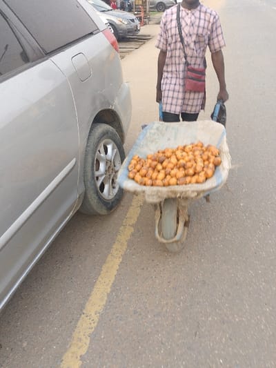 The image depicts a street scene, likely in Ijaiye, Nigeria, featuring a person pushing a wheelbarrow laden with produce alongside a parked vehicle. A person, dressed in a light-colored plaid tunic-style shirt and dark shorts, walks behind and slightly to the left of a blue wheelbarrow. A red shoulder bag is worn across their body, and they hold a dark object in their left hand.
The wheelbarrow, which appears well-used, contains a large pile of small, round, orange-brown fruits, possibly African star apples or a similar local fruit. The single wheel of the wheelbarrow is visible at the bottom center.
To the left, the rear side of a silver or light grey minivan or SUV is parked. The vehicle shows some scuff marks or scratches on its body panel. The scene is set on an asphalt road with a faded yellow line marking part of the lane. In the blurred background, other cars and what appear to be informal structures or market stalls suggest an urban or semi-urban environment. The lighting is soft and diffused, indicating daytime, possibly under an overcast sky. No visible text is legible in the image.
