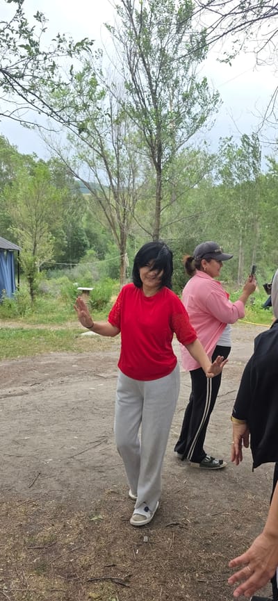 In Кашка-Суу, Kyrgyzstan, a woman with dark hair and bangs, wearing a red t-shirt with lace sleeves and light gray sweatpants, stands on a dirt path with her right arm extended and her left arm slightly bent, her hand in a waving gesture. She is smiling. Behind her, to the right, another woman in a pink shirt and a dark baseball cap is holding a smartphone and looking down at it. Further to the right, a portion of a person in a black jacket is visible. The background features a lush green environment with trees, foliage, and a cloudy sky. To the left, a small blue structure with a pitched roof and draped blue fabric is partially visible. The ground is covered with dirt and scattered dry twigs.