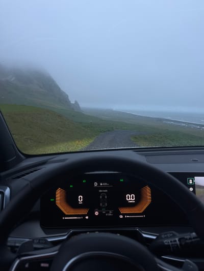 The image shows a view from inside a car, likely a hybrid or electric vehicle, driving through a landscape in Vik, Iceland. The dashboard is visible, displaying a speedometer reading of 0 km/h and a temperature of 8°C. The car appears to be in "D" (drive) mode, with "EV" and "ECO" indicators lit up. The mileage of the car is 6139 km.

Outside the car, the weather is overcast and foggy, obscuring the view of the coastline and grassy hills. A narrow road is visible winding through the landscape. The overall atmosphere is one of a misty, somewhat desolate environment.