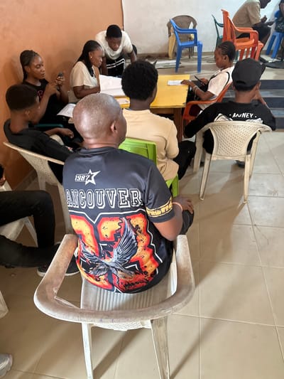 This image depicts an indoor gathering of approximately ten individuals in the city of Iriebe, Nigeria. The scene is observed from a slightly elevated perspective behind a bald man in the immediate foreground, who is seated on a cream-colored plastic chair with his back to the camera. He wears a black short-sleeved shirt featuring "1987 VANCOUVER" and an eagle graphic.

To the right of the foreground man, another person is seated on a green plastic chair, seen from behind, wearing a beige shirt. Further to the right, a third individual, also seen from behind, wears a black long-sleeved shirt with "MONEY" printed on the back and a black baseball cap, seated on a white plastic chair.

In the mid-ground, a rectangular yellow table serves as a central point for four individuals. On the left side of the table, a young woman in a black t-shirt holds a mobile phone, looking towards the right. Next to her, a young woman in a beige t-shirt examines papers on the table. Directly across from her, a young man in a light-colored shirt leans over the table, also focused on papers, seemingly interacting with the woman in beige. To the right of the table, a young woman in a white t-shirt looks down at a mobile phone.

The environment features light brown-orange walls and a floor tiled with large, light-colored square tiles. Various plastic chairs are scattered throughout the room, including blue and orange chairs visible in the background, where additional indistinct figures are present. A raised platform or step is also visible in the background on the right. The overall activity suggests a meeting, study session, or group discussion.