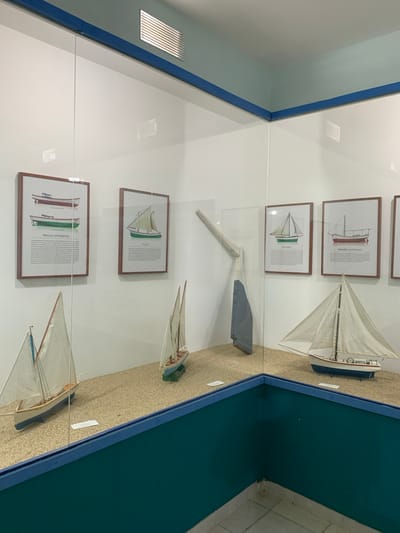 The image shows a museum display, likely in Boca del Río, Venezuela, featuring model boats and informational posters behind glass. The display area appears to be a corner with white walls and a blue trim along the top. The floor is tiled. The display itself has a sandy surface where the models are placed.

In the foreground, to the left, is a model of a sailboat with white sails and a blue and white hull. To the right of this is another model of a sailboat with white sails and a wooden hull, resting on a green base. Further to the right, another white sailboat model with sails is positioned on the sandy surface. Behind these models, on the wall, are framed posters. The posters are images of different types of boats, each with accompanying text. One poster on the left depicts three styles of boats, with the title "PIRAGUA MOTORIZADA". Another poster to its right shows a sailboat with the title "BALICHORO". To the right of that, another poster shows a sailboat titled "TRES PUNTO". A large oar-like object, painted in light wood and blue, is mounted on the wall to the left of the posters. The overall impression is an exhibit dedicated to maritime history or local seafaring traditions. There are no people visible in the image.