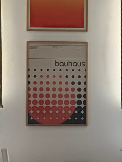 The image shows a wall with two framed posters. The poster on the left has an orange gradient. The poster on the right features the word "bauhaus" vertically aligned on the left side. To the right of the text is a pattern of circles, starting with black circles on the upper right and gradually transitioning to red circles, forming a semi-circular shape on the lower right. Below the text, the poster also contains the text "staatliches bauhaus, 100th anniversary, german art school", "walter gropius, weimar, dessau, berlin, school of architecture" and "modernism, minimalism & abstract arts". The location is Clichy, France.