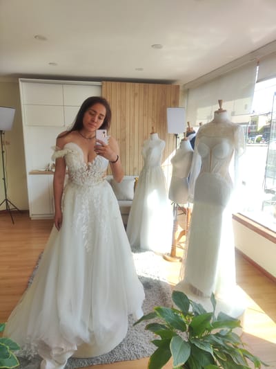 The image shows a woman taking a selfie in what appears to be a bridal shop. The woman is wearing a white off-the-shoulder wedding dress with floral lace detailing on the bodice and a flowing skirt. She is holding a smartphone with a light purple case.
In the background, there are two mannequins wearing wedding dresses. One is a classic A-line gown and the other is a more modern, form-fitting dress with sheer sleeves and intricate beading. A third mannequin, draped with fabric, is also visible. The shop has a neutral color palette with white cabinets, light wood paneling, and a gray rug. There is a plant with large green leaves in the foreground. Natural light streams in through a large window on the right side of the image. The location is likely a retail space in Tacna, Peru, but there are no external identifiers visible in the image to confirm this.