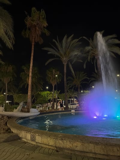This is a nighttime outdoor shot of a fountain in Sant Antoni de Portmany, Spain. The fountain is illuminated with blue and purple lights, with water spraying upwards. Palm trees surround the fountain, and a few people are visible in the background near a white car. The area appears to be a promenade or public square with paved ground and a stone border around the fountain.