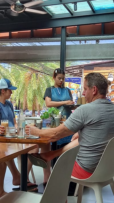 The image shows the interior of an outdoor restaurant in Ban Ao Nang, Thailand. A fair-skinned man in a gray T-shirt and red shorts leans over a wooden table, engaging with a woman with dark hair wearing a denim overall dress. The woman holds a pen. On the table are several empty glass bottles and glasses. Behind the woman is a street scene with lush green trees, power lines, and storefronts; a sign for "The Signature" is visible in the background. Another person wearing a blue baseball cap and a blue shirt is seated on the other side of the table. Feet and sandals can be seen beneath the table. A ceiling fan is visible in the upper left corner of the image.