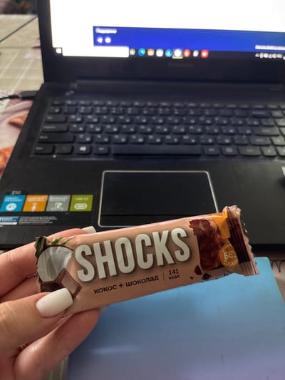 The image shows a person's hand holding a chocolate bar over a laptop keyboard. The chocolate bar has a light brown wrapper with the brand name "SHOCKS" printed in large, bold, white letters. Below the brand name, it says "КОКОС + ШОКОЛАД" which translates to "COCONUT + CHOCOLATE" in Russian. There is also text that appears to be "141 ккал," indicating 141 calories. The wrapper also features an image of coconut and a piece of chocolate. The laptop appears to be open, with its screen displaying some content, though it is mostly out of focus. The setting is likely indoors, on a desk, as suggested by the laptop and the presence of a light-colored object (possibly a notebook or book) next to it. The lighting is somewhat dim, suggesting it might be evening or a dimly lit room. The overall impression is a casual scene of someone taking a break or snacking while working or browsing on their laptop. There are no strong emotional cues evident, and the weather and time of day are not clearly discernible.