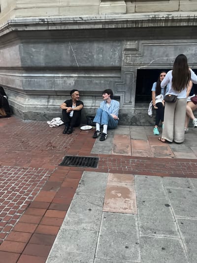 The image shows a group of people on a paved street in what appears to be an urban setting. The scene is oriented vertically, suggesting the photo was taken with a phone held sideways.

In the foreground, a man in a black t-shirt and black pants sits on the red brick pavement, facing left with his hands clasped. A white garment is folded on the ground near him. Another man, wearing a blue and white striped shirt and blue jeans, sits on the grey stone pavement to his left, also facing left and appearing to be looking down.

Further back and to the left, a group of people are partially visible. One person with long brown hair is wearing a white shirt and light-colored pants, and is sitting down. Another person is wearing a light blue shirt. Some of these individuals appear to be leaning against a large, dark architectural structure with decorative molding.

The ground is paved with a combination of red brick and grey stone blocks. A metal grate is visible on the red brick portion of the pavement. The architecture in the background suggests a building with stone facades.

The lighting indicates it is daytime, possibly late afternoon, given the shadows. There are no overt cues for weather. The overall impression is of people casually gathered or resting on a street. The location context provided is Bilbao, Spain, which aligns with the architectural style and paving materials often found in European cities. No visible text is present in the image.