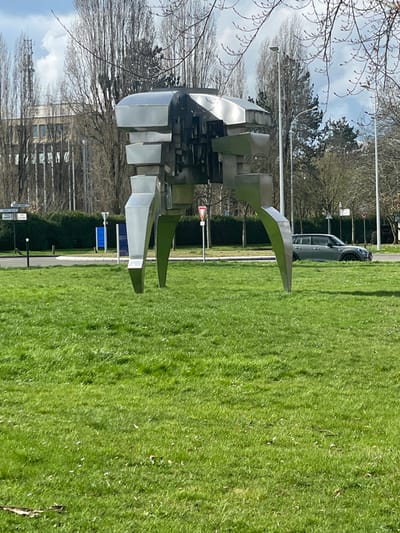 The image displays an outdoor urban environment in Montigny-le-Bretonneux, France, under natural daylight. A large, abstract, bipedal metallic sculpture dominates the central frame, positioned on a vibrant green lawn. The sculpture is constructed from angular, reflective stainless steel panels, forming a complex, segmented body with two prominent, splayed legs in the foreground and two in the background. The immediate foreground features bright green grass, flecked with small white or yellow flowers.
Behind the sculpture, a paved road or driveway is visible. A dark grey four-door Mini Countryman car is parked facing right on the roadside, partially obscured by a low dark green hedge and one of the sculpture's legs. To the left of the sculpture, a red-bordered white triangular "Cédez le passage" (Yield) road sign is present, alongside a blue rectangular sign and two streetlights. The background features a dense line of tall, bare-branched deciduous trees, suggesting late winter or early spring. Beyond these trees on the far left, a multi-story, light-colored building with visible windows stands, topped by a slender telecommunications tower. The sky above is light blue with scattered white clouds. No individuals are visible.