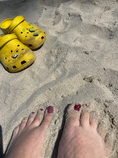 This is a close-up, overhead shot of a person's feet partially buried in sand, with two yellow Crocs shoes resting on the sand to the upper left. The feet have dark red glitter nail polish on the toenails and are covered in fine grains of sand. The Crocs shoes also appear to have some sand on them and are adorned with colorful jibbitz charms. The background is entirely composed of beach sand, with subtle shadows suggesting daylight. The location is identified as Kulikovo, Russia.