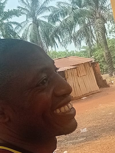 The image captures an outdoor scene in Ayangba, Nigeria, featuring a man in the foreground and a rustic structure with vegetation in the background. The man, with dark complexion, is depicted in a right-facing profile view, encompassing his head, neck, and upper shoulder. He has an open mouth with visible white teeth and upturned lips, consistent with smiling or laughing. His left eye is partially visible, and he wears a garment with red and yellow sections visible at the bottom left.
In the mid-ground, a small, weathered building is visible. It features a reddish-brown, rusty corrugated metal roof and walls constructed from vertical wooden slats or similar material. A darker, possibly brick or stone, section adjoins the wooden wall on its right side. The building stands on reddish-brown dirt ground, where several small, light-colored objects are discernible near its base. The background consists of multiple tall palm trees with green fronds, interspersed with other green foliage, set against a bright, hazy sky.