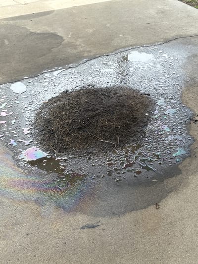 An aerial view of a pile of dark soil sits on a cracked concrete surface. A puddle of water surrounds the soil and exhibits an iridescent sheen, indicating the presence of oil. The scene is located in Ruskin, Florida, United States. The concrete shows signs of wear and tear, with a visible crack running diagonally across the frame. There are no people or animals present in the image. The overall impression is one of neglect or a minor spill.