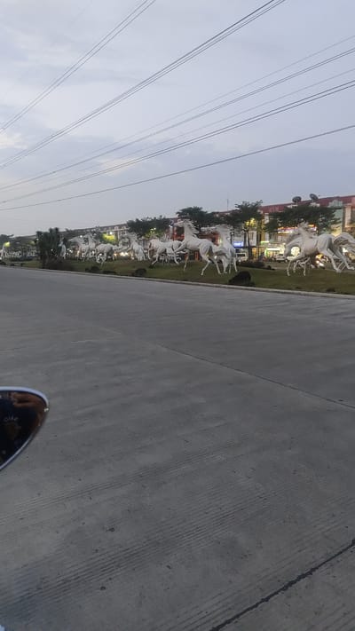 This media file captures a scenic view of multiple white horse sculptures arranged along a grassy median strip bordering a road. The sculptures are depicted in mid-gallop, creating a dynamic and majestic display. Behind the sculptures, a row of buildings with a mix of commercial and residential facades is visible, adorned with trees and streetlights. The sky above is overcast with a muted gray hue, suggesting either early morning or late evening. Power lines stretch across the upper portion of the frame, indicating an urban or suburban setting. The foreground shows a gray asphalt road, with a reflection of what appears to be a motorcycle's side mirror visible in the bottom left corner. The overall mood is serene and perhaps a bit tranquil due to the subdued lighting. There is no visible text in the image. The location context is Pusakarakyat, Indonesia.