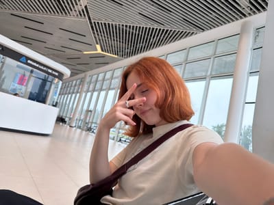 A medium shot shows a young, fair-skinned person with auburn hair sitting in what appears to be an airport terminal. The person is wearing a light-colored t-shirt and a crossbody bag with a dark brown strap. They are holding their left hand up to their face, fingers forming a "V" sign, with a ring visible on one finger. The background features a bank, TBC Bank, written in Georgian. Large windows let in daylight, revealing an overcast sky and trees outside. The architectural style of the building includes a modern ceiling with linear patterns and large white support columns. The floor is tiled. The location is Ianeti, Georgia.