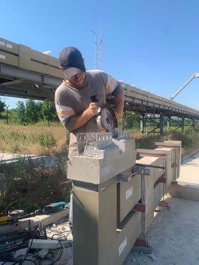 A man wearing a gray t-shirt with orange and white stripes and a dark blue baseball cap is using a circular saw to cut a concrete structure. Sparks are flying from the saw as it makes contact with the material. The man is focused on his work and appears to be wearing glasses. The concrete structure being cut is a small, intricate model, possibly of a building, with decorative elements. Several large concrete blocks are stacked behind the model, secured with metal brackets. In the foreground, on the ground, are various tools, including a yellow and black angle grinder and a teal-colored rotary hammer drill, along with electrical cords and debris. The scene is outdoors, with a bright blue sky overhead and lush green vegetation in the background, including some red wildflowers. A long, elevated structure, likely part of an industrial or transportation facility, runs across the background. The location is identified as Pankratove, Ukraine.