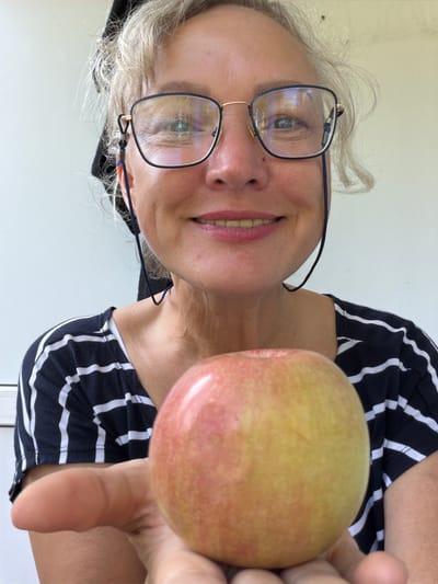 In Shepsi, Russia, a woman with blonde hair pulled back and wearing glasses and a black and white striped shirt is holding an apple. The woman is smiling and looking at the camera. The background is a plain white wall.