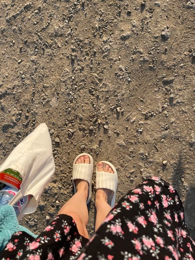 This media file shows a close-up, overhead view of a person's legs and feet on a gravelly path, with a black floral dress and white slides visible. A light-colored tote bag rests on the ground beside their feet, containing a bottle of water and other items. The scene suggests a casual outing in a dry, outdoor environment. The lighting indicates it is likely daytime, possibly late afternoon or early morning due to the long shadows cast by the gravel. The setting appears to be a path or unpaved area, consistent with a rural or coastal location like Ahtopol, Bulgaria, as suggested. There is visible text on a label within the tote bag, which reads in part: "тел: 0897 02 06 28" and "email: gpobel@gmail.com". The overall mood is relaxed and unhurried.