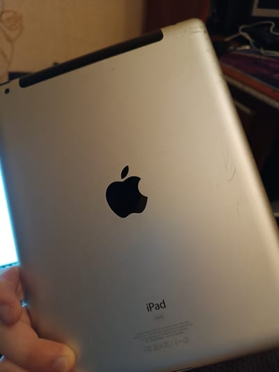 The image shows the back of a silver iPad. The Apple logo is visible in the center, and the word "iPad" with "64GB" below it is printed at the bottom. There are several scratches on the back of the iPad, particularly near the top right corner and to the right of the Apple logo. A person's hand is holding the bottom left edge of the iPad. The background is blurry and appears to be an indoor setting with a wall and some objects on a shelf or desk. There is no identifiable information in the image that places it in the city of Kozmodemyansk, Russia.