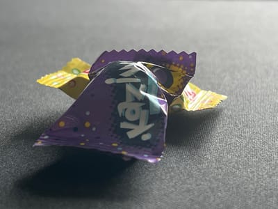 The image shows three candy wrappers on a dark, textured surface. The wrapper in the foreground is purple with white lettering and yellow and blue dots. The lettering appears to be in a stylized font and resembles the word "KAZ". Behind it, two yellow wrappers are partially visible, one with colorful stripes and circles, and the other with similar colorful markings. The lighting creates shadows on the surface, suggesting a single light source. There are no people or animals in the image, and no context is provided regarding the location, including the city of Kalininets, Russia.