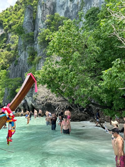 The image depicts a vibrant scene at Ton Sai Beach, Thailand, characterized by its clear turquoise waters and towering limestone cliffs. Several tourists are seen wading in the shallow, clear water, some appearing to disembark from a traditional longtail boat, indicated by its prominent wooden prow adorned with colorful garlands. The beach is backed by lush green vegetation cascading down the steep, rocky cliffs. People are dressed in swimwear, with some holding phones, suggesting they are capturing the scenery or interacting. The overall atmosphere is one of a popular tourist destination, bustling with activity and bathed in natural light under a bright sky.