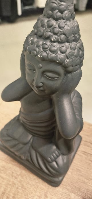 A dark grey, matte-finished statue of a Buddha-like figure is depicted seated in a meditative posture, approximately 20-30 centimeters in height. The figure's hands are raised, with palms covering the ears, and its eyes are closed. A small, circular urna mark is present on the forehead. The head features a prominent ushnisha and sculpted curls, with multiple small white scuffs or paint chips visible on the top and side surfaces. The statue is positioned on a light brown wooden surface exhibiting visible grain. The background is out of focus, revealing a lighter-toned wall or surface behind the statue and an indistinct, dark object partially visible in the upper left section. The scene is located indoors in Vendin-le-Vieil, France.