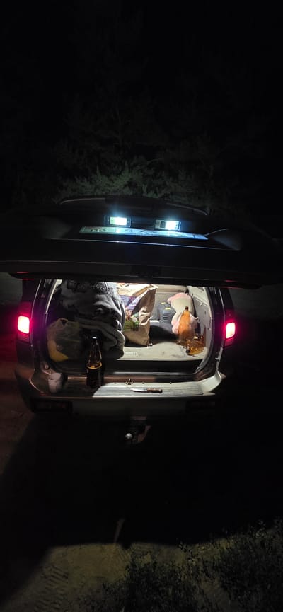 The image shows the open trunk of a car at night. The car is parked in an outdoor, unlit area, possibly a rural or undeveloped location, with trees visible in the background. The trunk contains several items, including bags of groceries, a blanket, plastic bottles, and a knife. The car's brake lights are illuminated, casting a red glow. The scene appears to be in the city of Karakol, Kyrgyzstan, though no specific landmarks or identifying features of the city are visible.