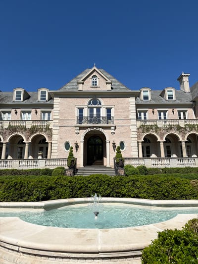 This image depicts a grand, multi-story building alongside a large water feature under a clear blue sky. The building, likely a stately home or institution, features light-colored brickwork and a multi-pitched roof with grey tiles. Its architectural style is ornate, possibly reflecting French Châteauesque or classical revival influences, with numerous windows, including rounded arches, rectangular panes, and prominent dormers on the roofline. Decorative elements include stone trim, columns, balconies with intricate wrought iron railings, and sconce lighting near the main arched entrance. Vines are seen climbing parts of the brick facade.
In the foreground, to the right of the building, a large, light-colored stone fountain or water feature contains clear, light blue water, with ripples and a small spray indicating a water jet. Manicured green hedges line the perimeter, separating the building from the fountain and adding to the cultivated landscape.
The scene is static, with no human activity or specific event taking place. The bright, clear blue sky suggests a sunny day, likely during the morning or afternoon, given the even lighting and lack of harsh shadows. The overall impression is one of grandeur, elegance, and tranquility. No visible text is discernible in the image. The setting is identified as Potomac, United States of America.