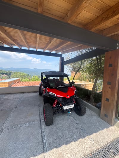 The image shows a red and white Z-Force side-by-side vehicle parked on a concrete surface with tire tracks, possibly a driveway or patio. The location is suggested to be Valle de Bravo, Mexico, based on the view of mountains and a landscape with red-tiled roofs visible in the background. The vehicle is positioned close to a building with an overhanging wooden roof supported by dark metal beams. A drainage grate runs along the edge of the concrete surface. There are no people visible in the frame.