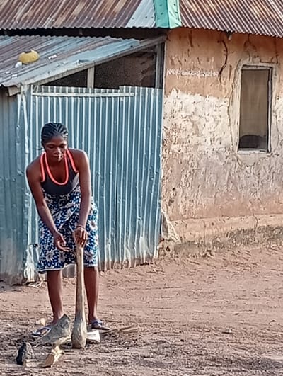 The image portrays a female individual performing an outdoor task on unpaved reddish-brown earth, within an environment consistent with a residential or semi-urban area of Gboko, Nigeria. No specific address or landmark within Gboko is identifiable from the image.
The subject, a dark-complexioned woman with braided hair, is depicted in a bent posture, facing downward. She wears a black tank top, an orange bra strap visible, a blue, white, and brown patterned skirt, and light blue flip-flops. Both hands grip a long, tapered wooden implement, resembling a pestle, which rests its wider end on the ground. Around the tool's base are scattered dark and light irregular fragments, consistent with wood, charcoal, or organic matter, including a larger, amorphous piece directly in front.
In the background, two structures are visible. To the left, a corrugated metal sheet wall stands beneath a sloped, rusty corrugated metal roof that includes a small light-green section and a yellow object. To the right, a building with an eroded, light-brown mud-plastered wall features a tall, narrow, unglazed window opening and faint white script. Its roof is also composed of rusty corrugated metal sheets.