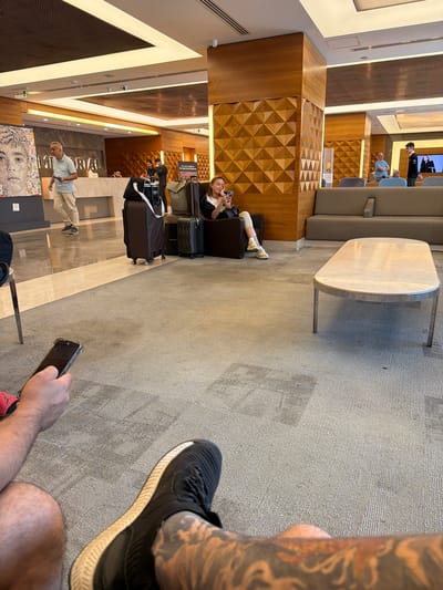 The image captures a hotel lobby in Şişli, Turkey. In the foreground, a person's legs and arm holding a smartphone are visible, suggesting they are seated. In the mid-ground, a woman sits in a dark armchair, looking at her phone. Several pieces of luggage are stacked near her. To the left, a man in a light blue polo shirt and beige pants walks by. Behind him, a reception desk is partially visible with a marble counter. The lobby features a modern design with wood paneling on columns and geometric wall decorations. A large marble-topped coffee table with a metal base sits in the center of the seating area, which includes a grey sofa. The ceiling has recessed lighting.