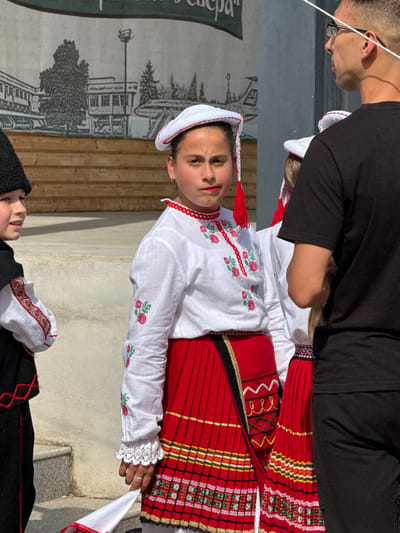 The photograph captures a cultural event in the city of Pordim, Bulgaria, likely a folk festival or performance. In the foreground, a young girl dressed in traditional Bulgarian attire stands prominently. Her costume consists of a white embroidered blouse with red floral motifs, a red pleated skirt with intricate woven patterns, and a white headdress adorned with red tassels. To her left, another child in a black and red traditional outfit is partially visible. Behind them, a man wearing glasses and a black t-shirt observes the scene. The backdrop features a wooden stage with a painted mural depicting buildings and trees. The overall atmosphere suggests a community gathering celebrating local heritage.