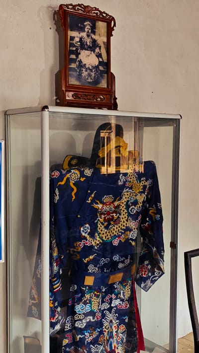 A glass display case contains a dark blue, intricately embroidered robe with a dragon motif. On top of the display case sits a framed photograph of a man in traditional clothing. The scene appears to be a museum setting, potentially in the city of Sà Phìn Commune, Vietnam, based on the style of the clothing and artifacts. The display case has a white frame. The framed photograph has a dark wooden frame with ornamental details. The wall behind the display is off-white. Part of a dark wooden chair is visible on the right side of the frame.